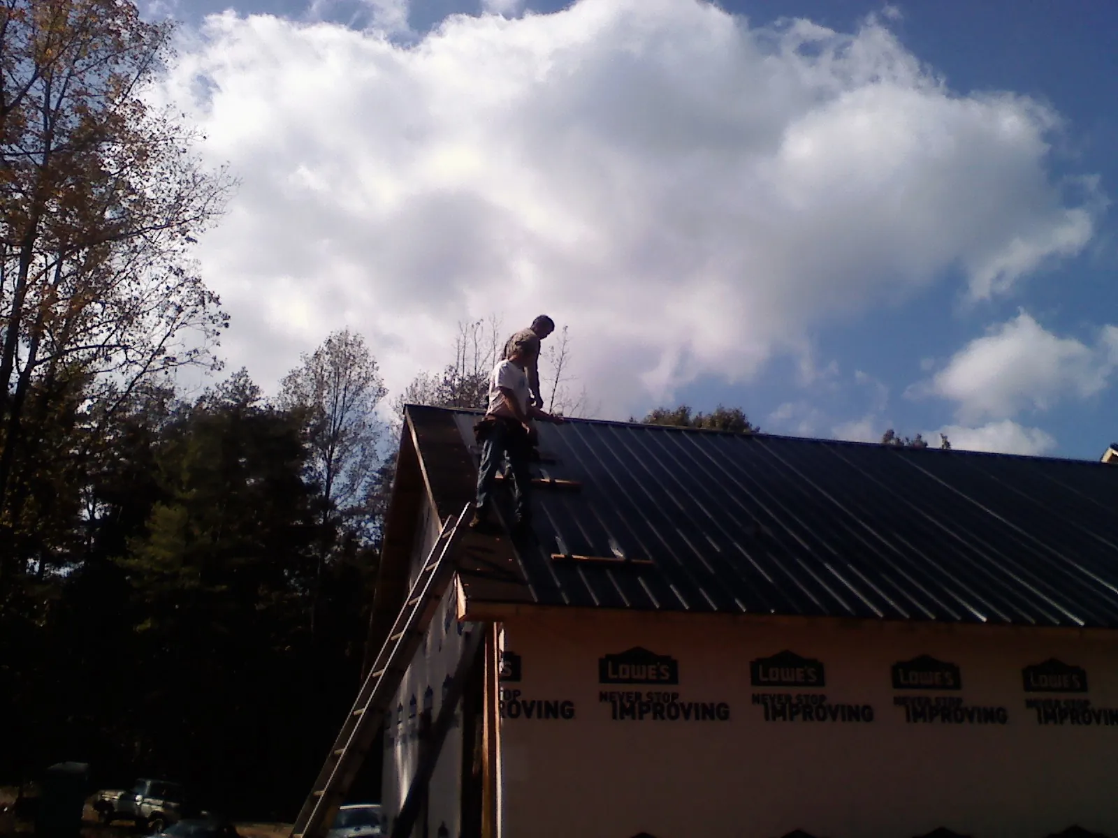 Roofer fastening dark metal panels high against a cloudy sky, from a Roof Inspection job in Alpha